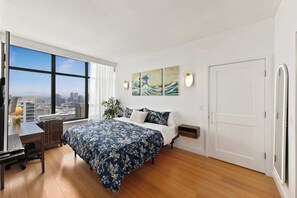 Ein Schlafzimmer mit einem großen Bett, einem Schreibtisch, einem Stuhl und Blick auf die Stadt durch große Fenster.