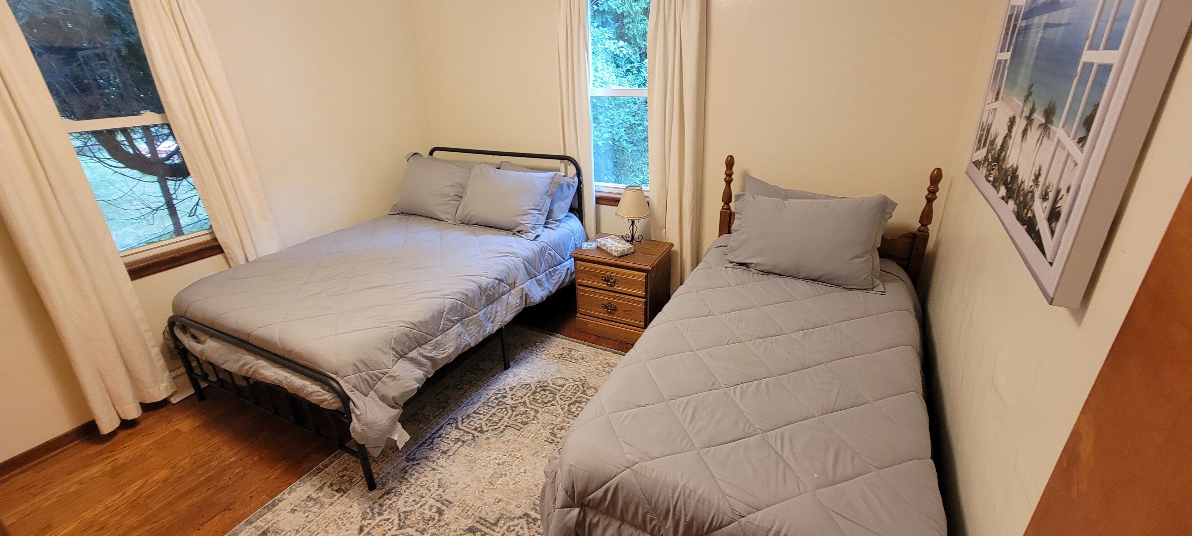 Un dormitorio con dos camas individuales, una mesita de noche con lámpara y una ventana con vistas a árboles.