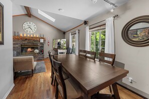 A dining area with a wooden table and chairs, a fireplace, a clock, and a large window with curtains. - Calm & Cozy Cabin - Cabin Retreat w/Spa & Large Fenced Yard (Big Bear City)