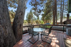 A wooden deck with a glass table and chairs, surrounded by trees. - Calm & Cozy Cabin - Cabin Retreat w/Spa & Large Fenced Yard (Big Bear City)