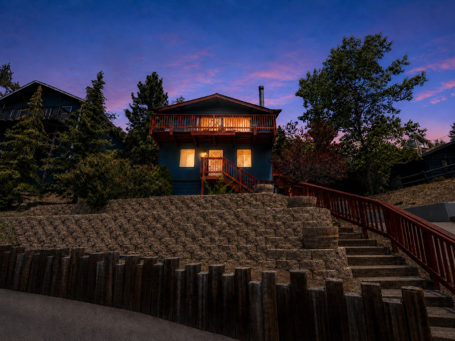 Une maison à deux étages avec un escalier en bois menant à l’entrée, entourée d’arbres et d’un mur de pierre.