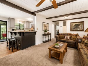 A spacious living area with a wooden coffee table, a leather sofa with patterned cushions, and a kitchen area with a bar counter and stools.
