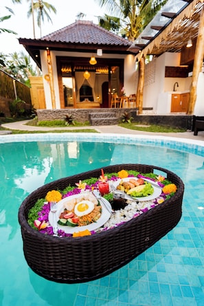 Disposición de comedor junto a la piscina, con una cesta de mimbre que contiene una comida, una piscina y un edificio con tejado de azulejos al fondo.