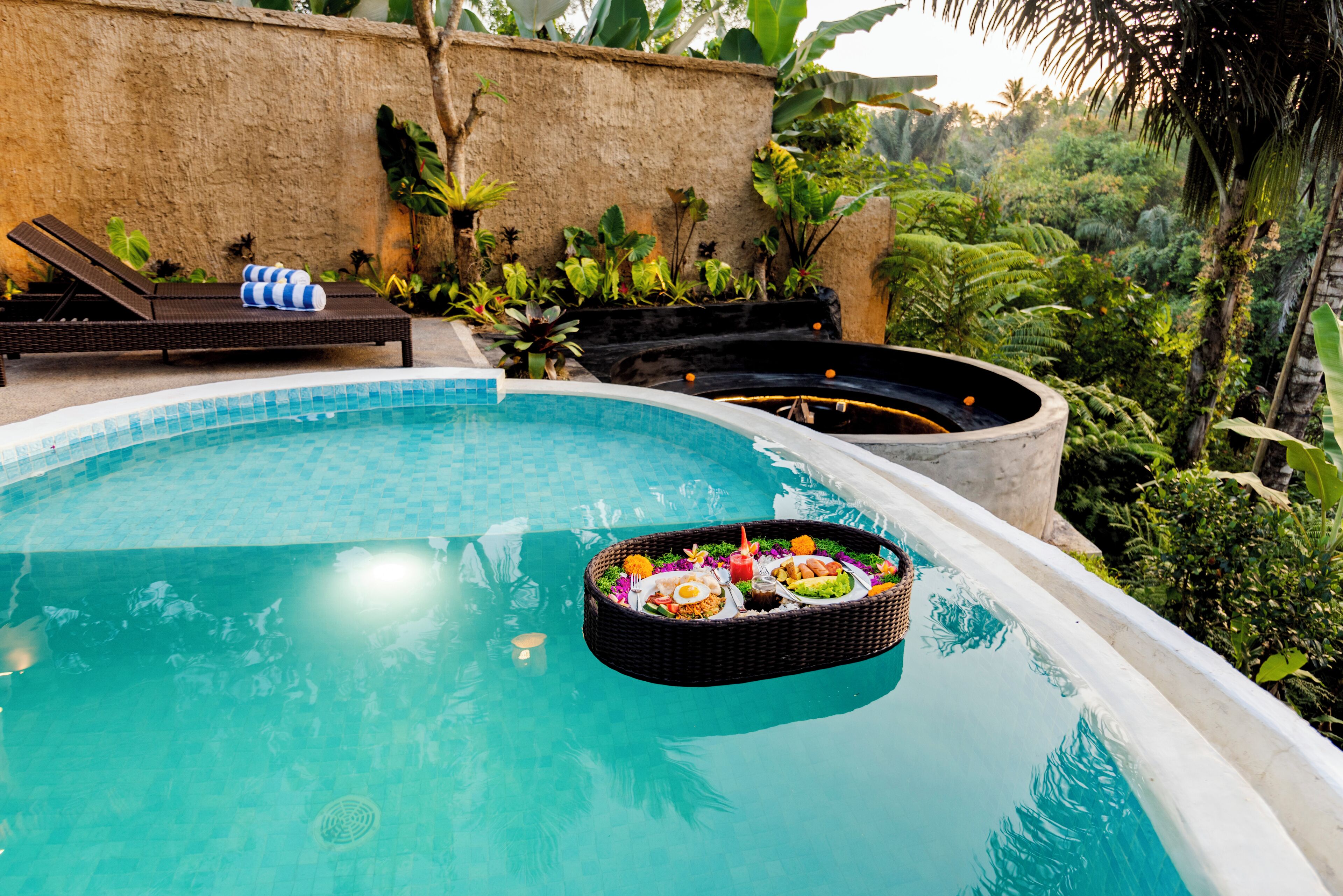 A swimming pool with a floating basket of food, lounge chairs, and tropical plants.