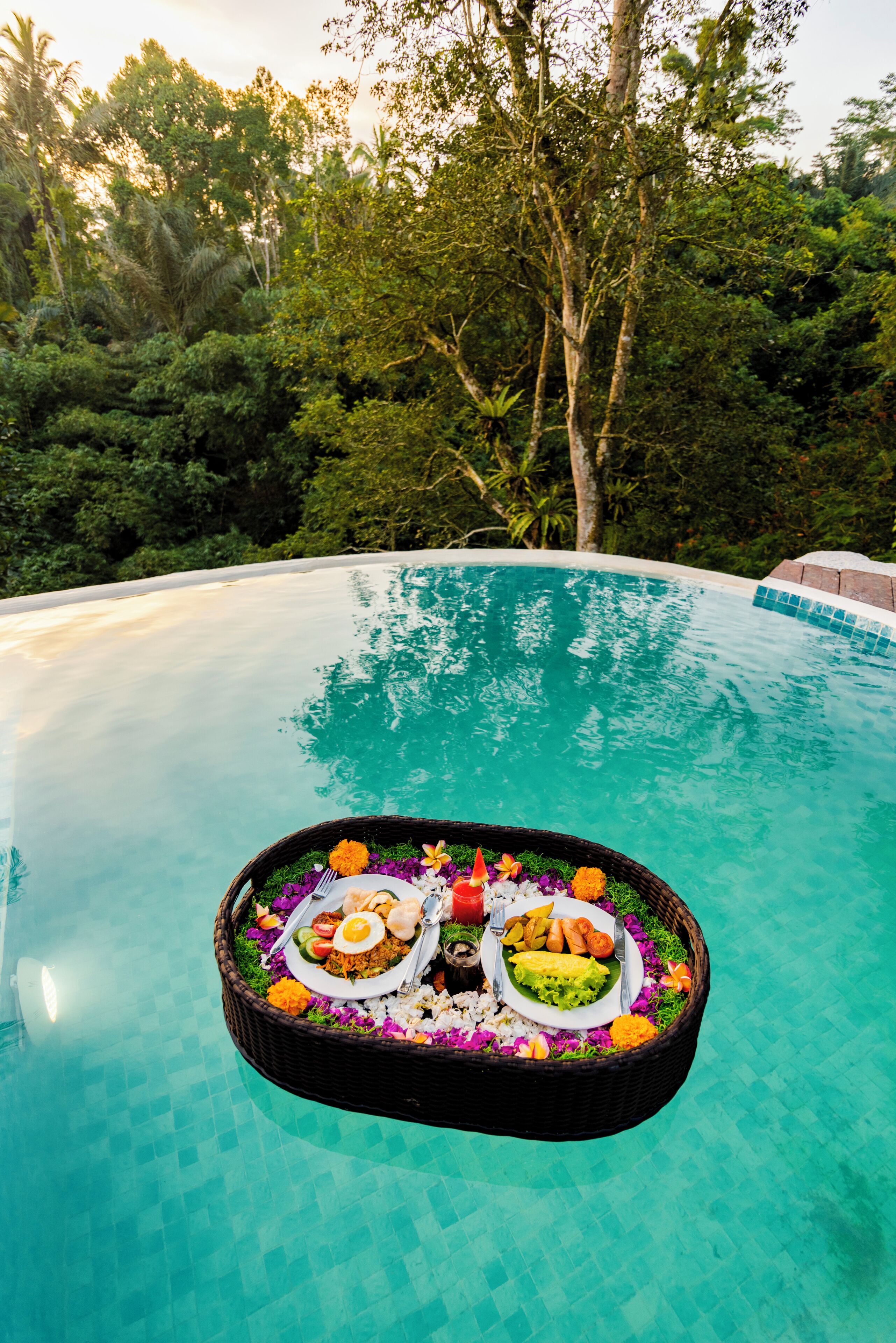 A poolside dining setup with a wicker basket containing a meal, surrounded by lush greenery.