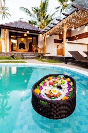 A poolside dining setup with a wicker basket containing a meal, surrounded by a pool with clear blue tiles. - Uma Harum Sari by Dhananjaya Hospitality (Gianyar)