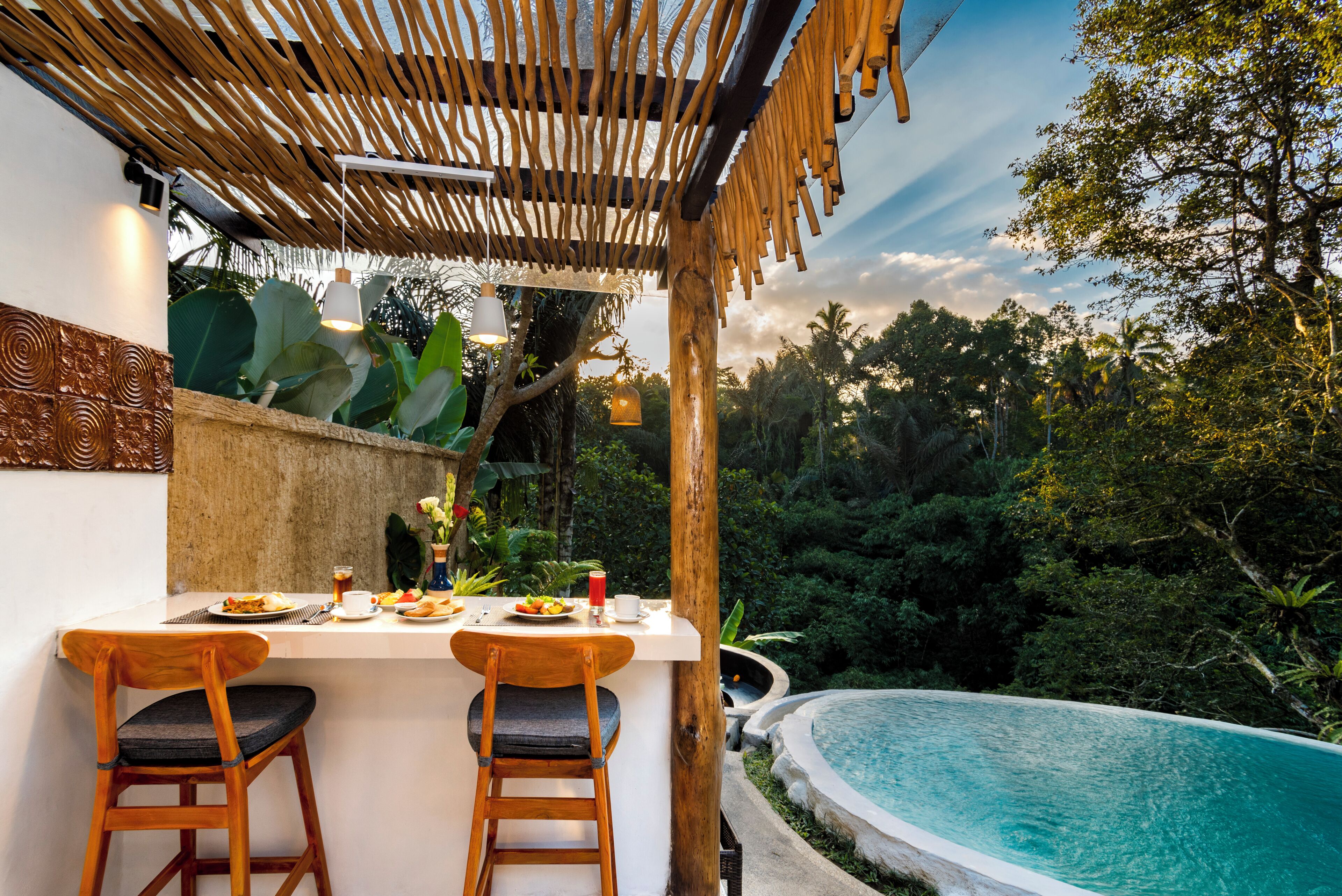 A dining area with a poolside view, featuring a wooden bar and chairs, a table set with plates and drinks, and a tropical backdrop.