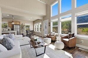 A spacious living area with a sofa, coffee table, and chairs, large windows with a view of the outdoors, and a kitchen in the background.