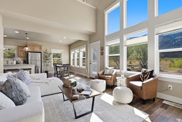 A spacious living area with a sofa, coffee table, and chairs, large windows with a view of the outdoors, and a kitchen in the background.