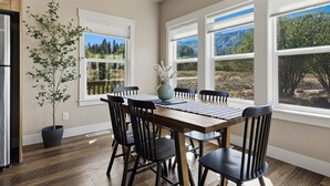 Un comedor con mesa y sillas de madera, ventanas amplias con vistas a árboles y montañas, y una planta en maceta.