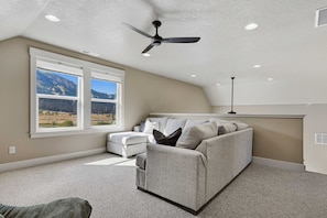 A spacious living room with a high ceiling, a ceiling fan, a large window with a mountain view, a grey sofa with cushions, and a carpeted floor.