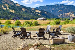 A lakeside outdoor seating area with Adirondack chairs and a fire pit, surrounded by a gravel ground and a view of mountains and a lake.