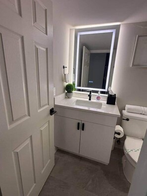 Ein Badezimmer mit weißem Waschtisch, einem Spiegel mit Beleuchtung unter dem Schrank, einer Toilette und einem kleinen Topfpflanzen.