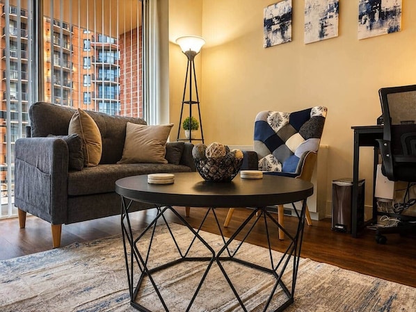 Ein modernes Wohnzimmer mit einer Couch, einem Couchtisch und einem Sessel. Eine Stehlampe und Kunstwerke sind an der Wand zu sehen.