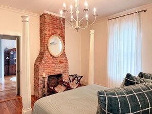 A bedroom with a brick fireplace, a chandelier, a bed with pillows, and a view of a hallway.