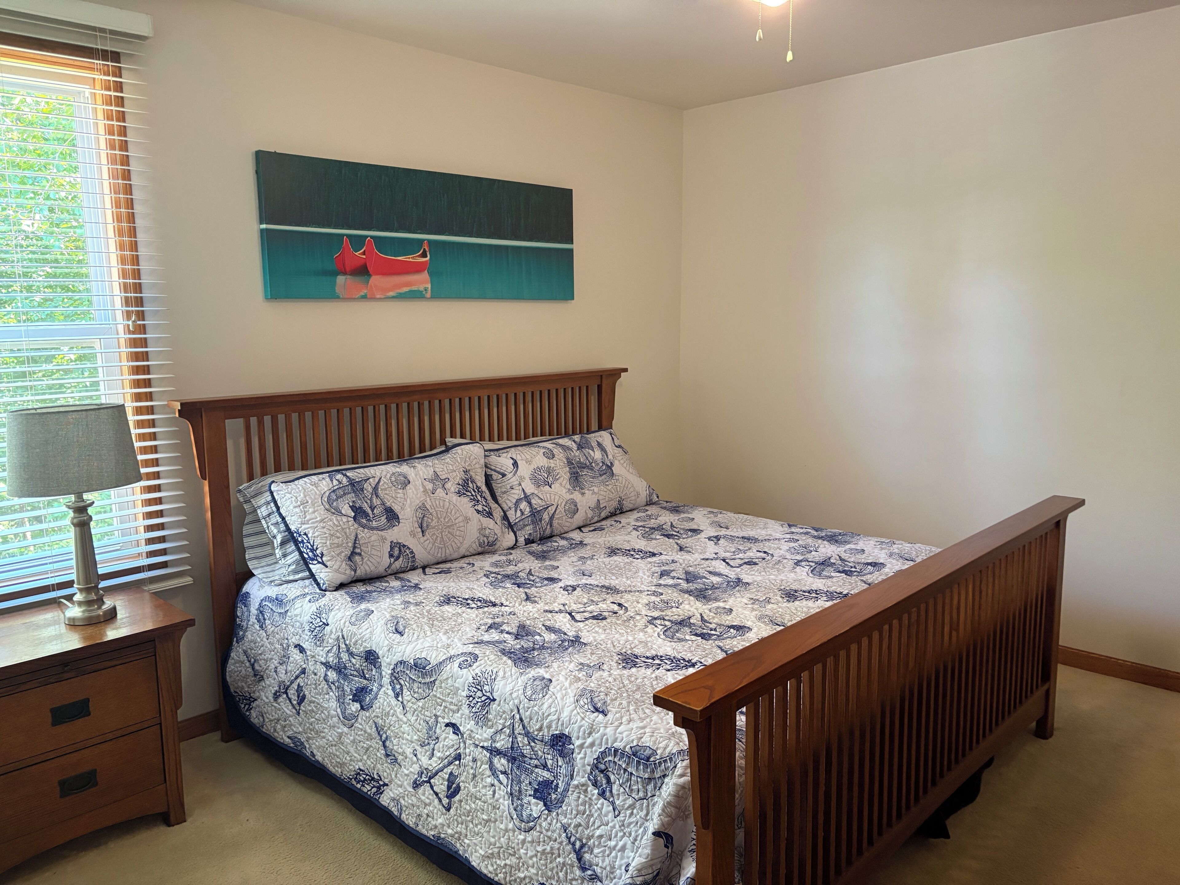 Un dormitorio con cama de madera, una mesita de noche con lámpara, una ventana con persianas y un cuadro colgado en la pared.