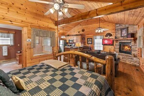 A cozy log cabin interior with a bed, a fireplace, a kitchen area, and a ceiling fan. - Cabin with WhirlpoolTub, Free Attraction Tickets, Fireplace and Gated Community! (Branson)