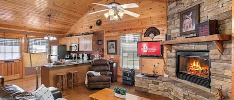 A cozy cabin interior with a stone fireplace, wooden ceiling, leather sofa, and a kitchen area.