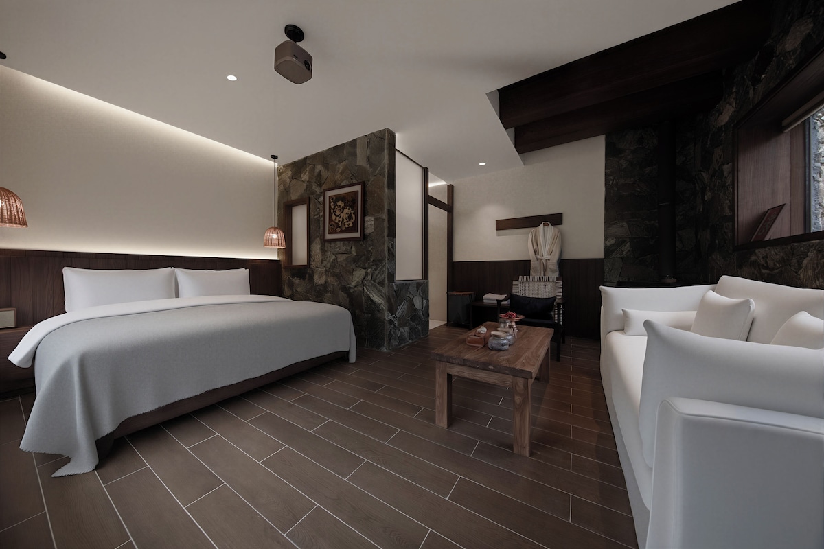 A modern hotel room with a large bed, a seating area with a table, and a stone accent wall.