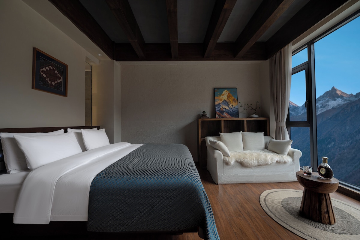 A bedroom with a bed, a sofa, a painting, a window with a mountain view, and a small table with a bottle.