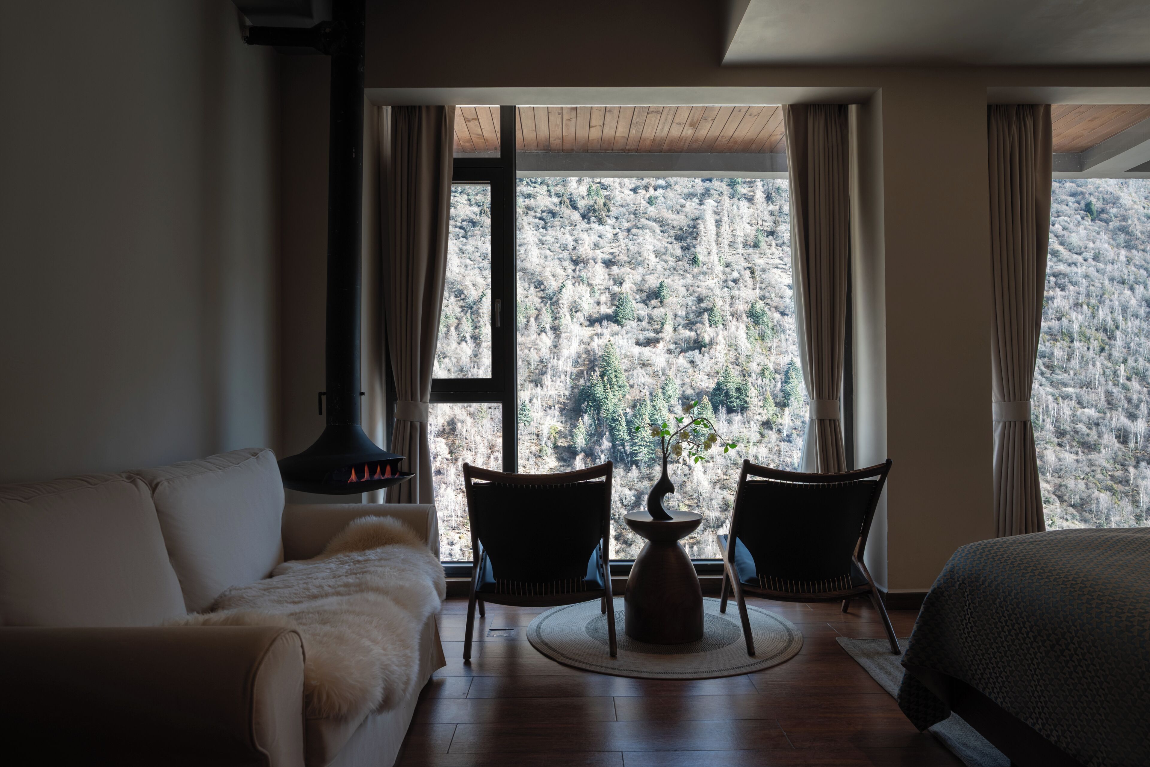 A room with a sofa, two chairs, a table with a vase, and a large window with a view of a forested hillside.