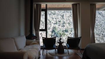 A room with a sofa, two chairs, a table with a vase, and a large window with a view of a forested hillside.
