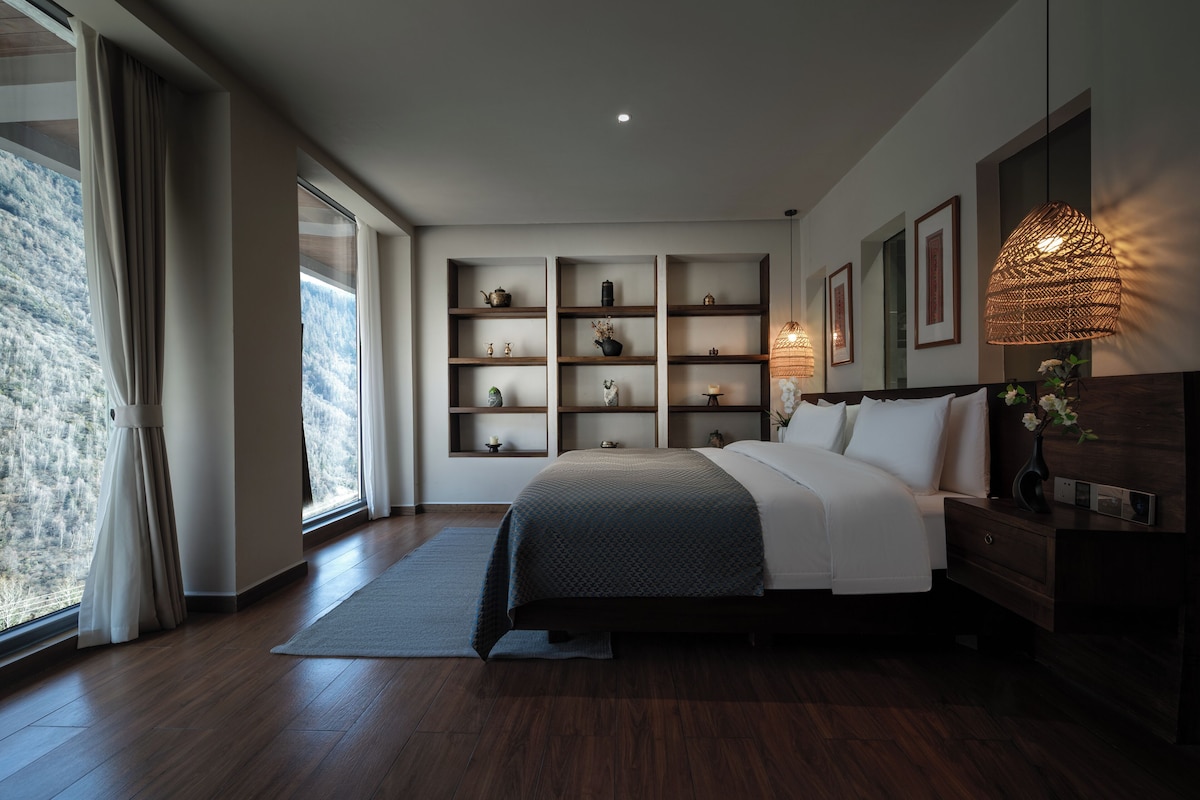 A modern bedroom with a large bed, wooden flooring, built-in shelving with decorative items, a large window with curtains, and a bedside table with a lamp.
