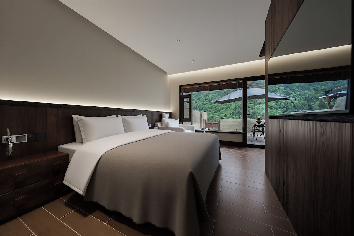 A modern bedroom with a large bed, a nightstand, and a view of a lush landscape through a large window.