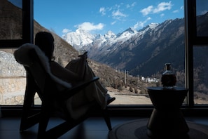 Une personne assise sur une chaise, devant une fenêtre d’où l’on peut admirer des montagnes enneigées.