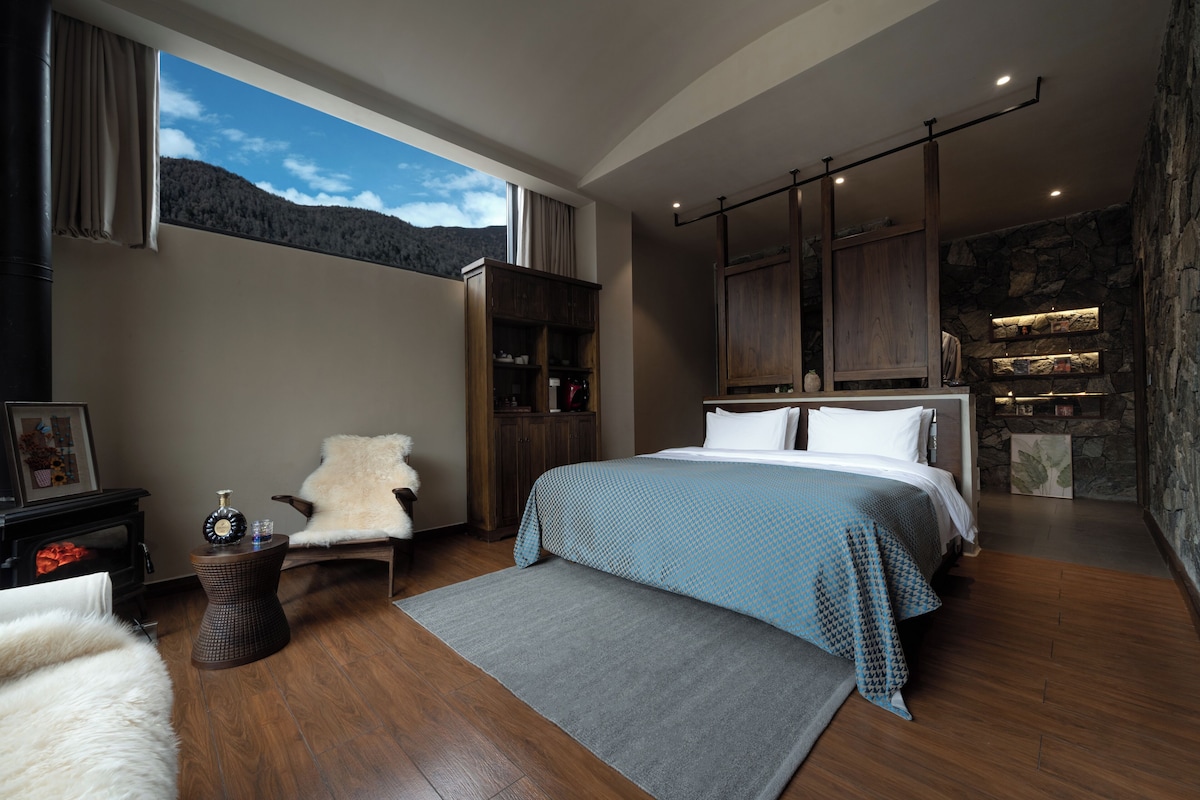 A modern bedroom with a large bed, a fireplace, a stone accent wall, and a view of the outdoors.