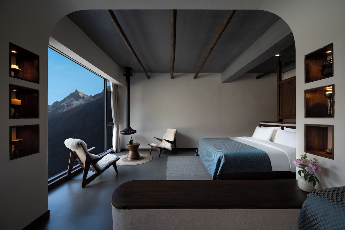 A modern bedroom with a large window, a fireplace, a bed, a chair, and a small table.