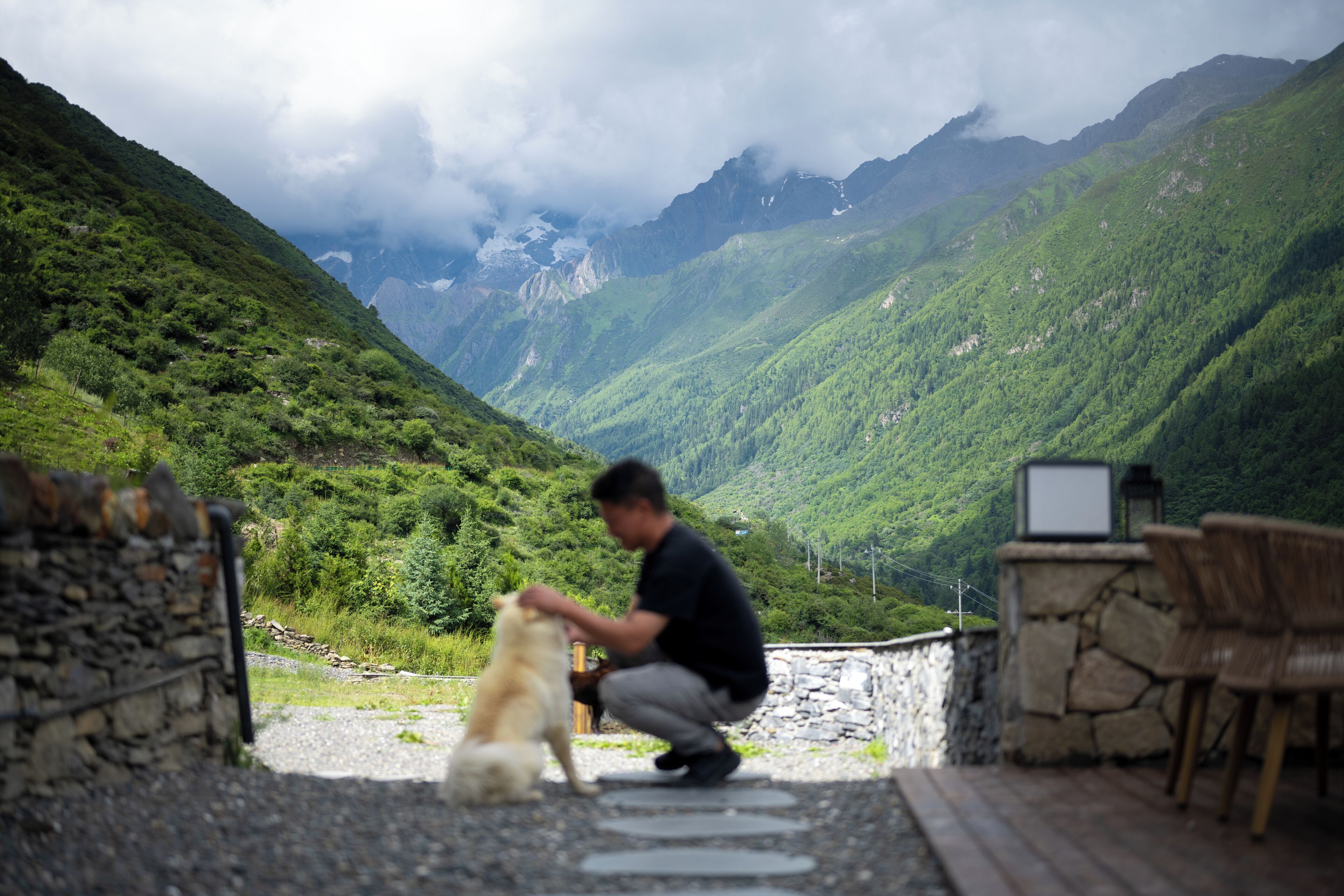 Una persona sentada en un banco de piedra al aire libre con un perro, con vistas a un paisaje montañoso.