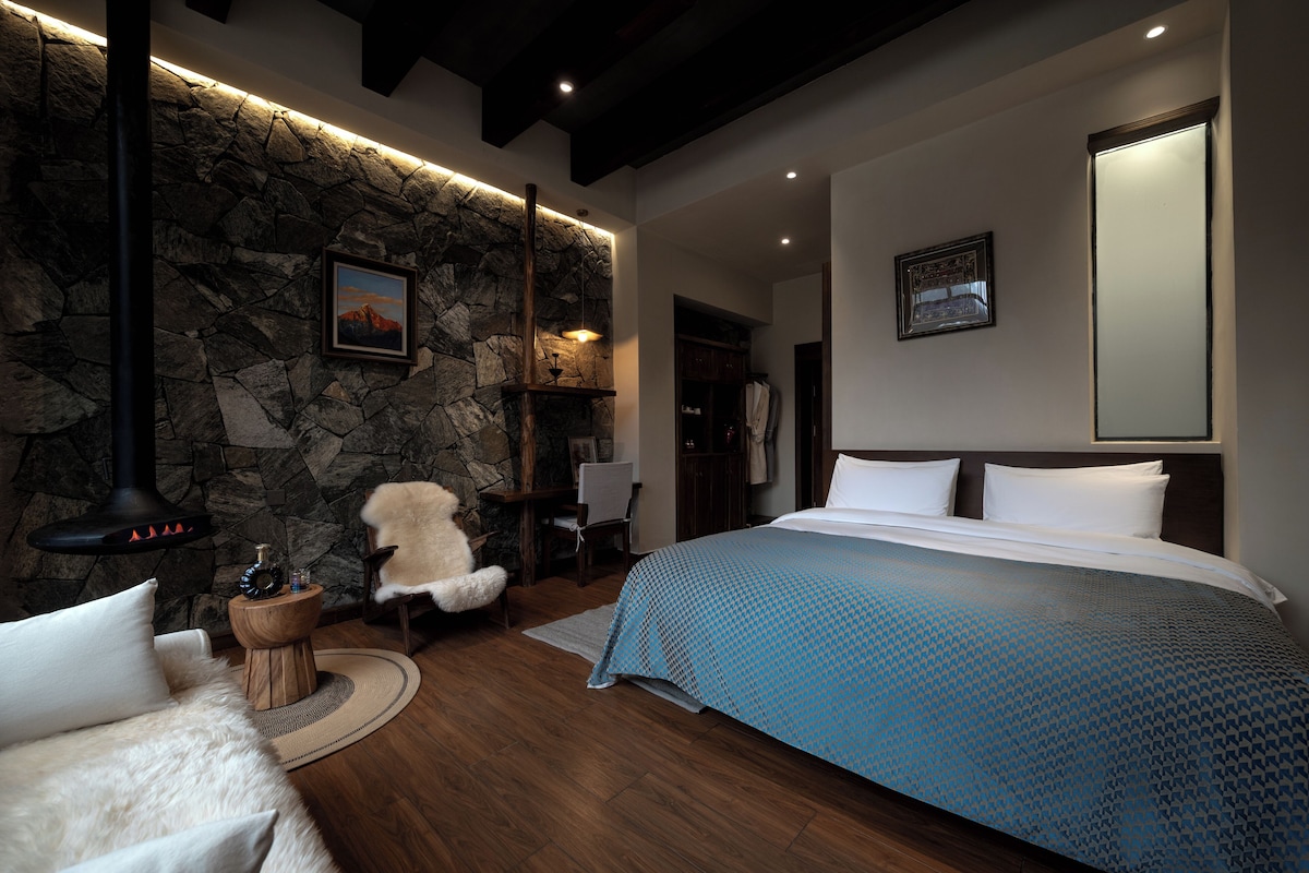 A bedroom with a stone wall, a bed with a patterned bedspread, a chair, a wooden side table, and a hanging light fixture.