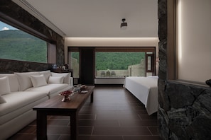 A modern hotel room with a stone accent wall, a large window overlooking a forested area, a sofa, a dining table, and a bed.
