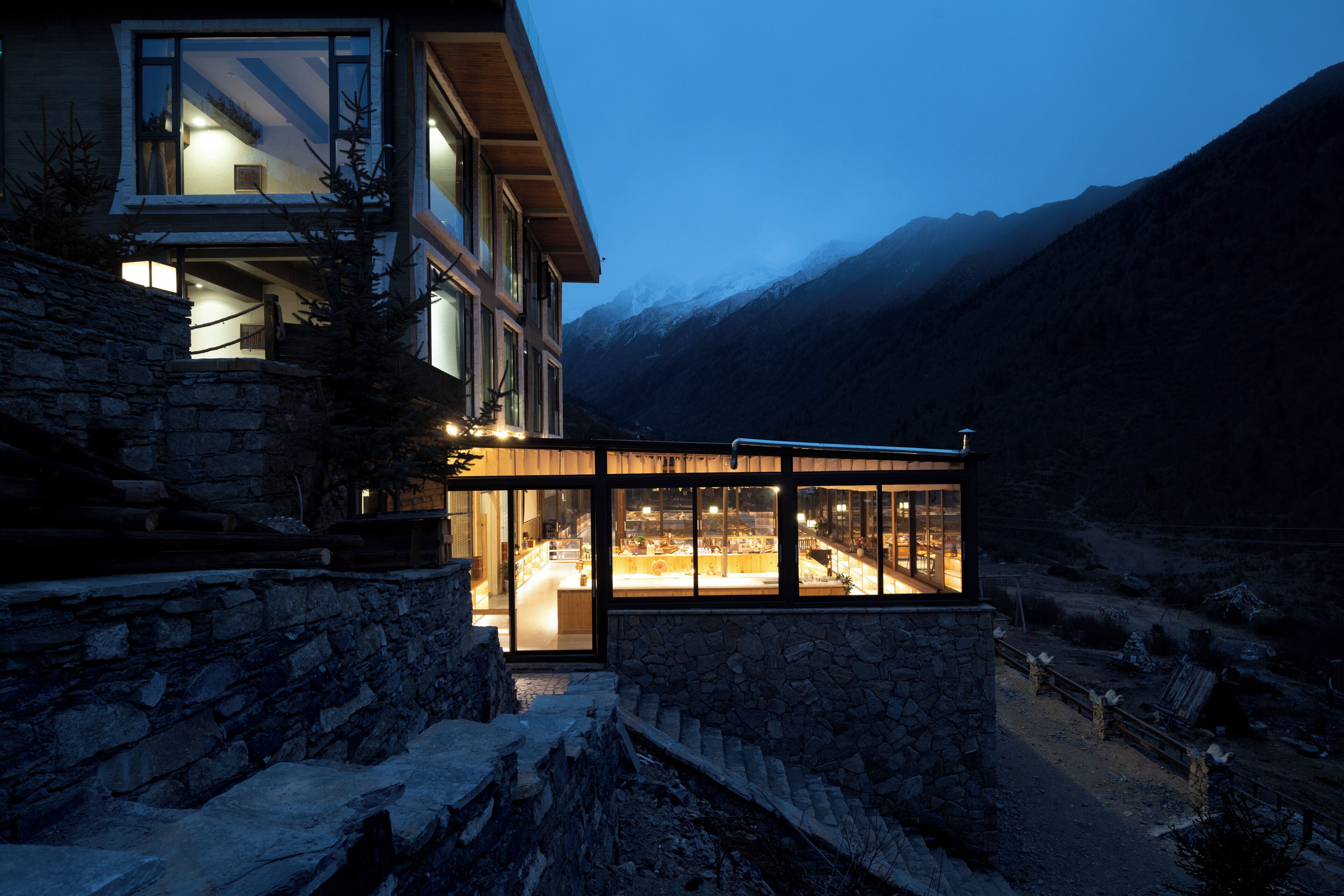 Une maison moderne avec une extension en verre, des murs en pierre et une vue sur les montagnes.