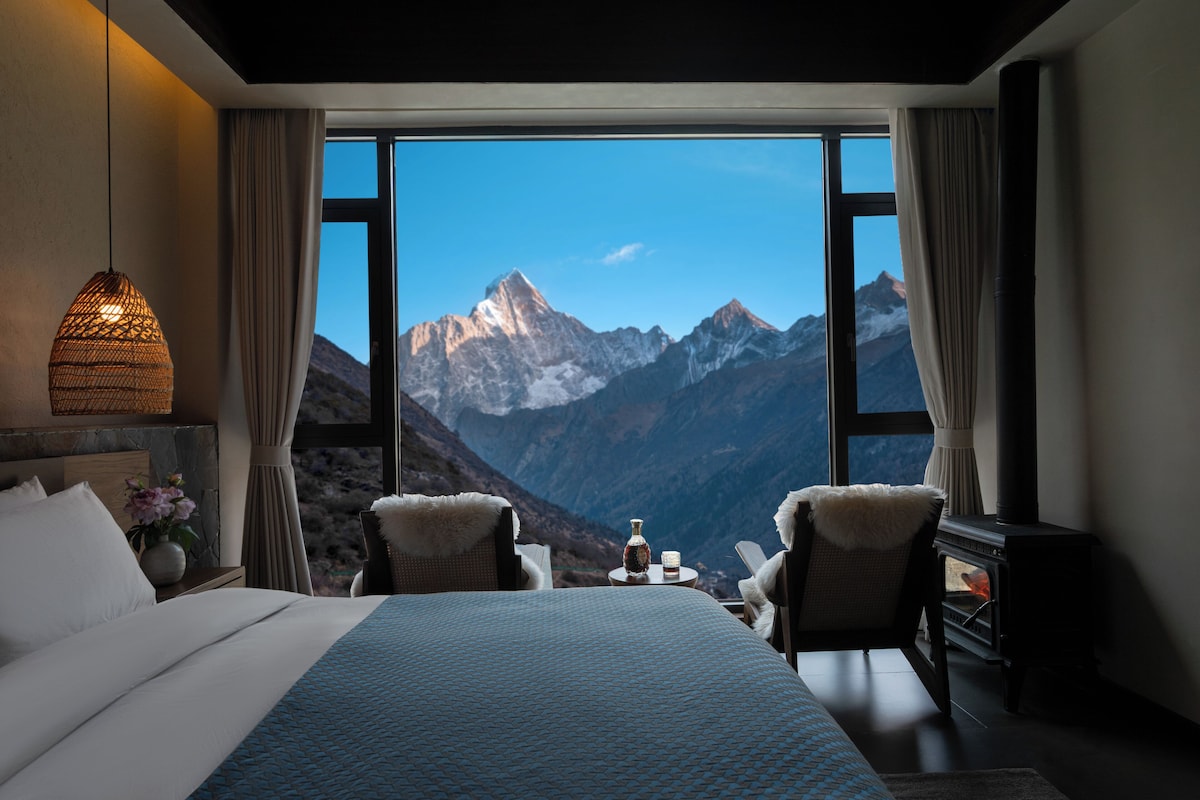 A hotel room with a large window offering a mountain view, a bed, a table with a bottle and glasses, and a fireplace.