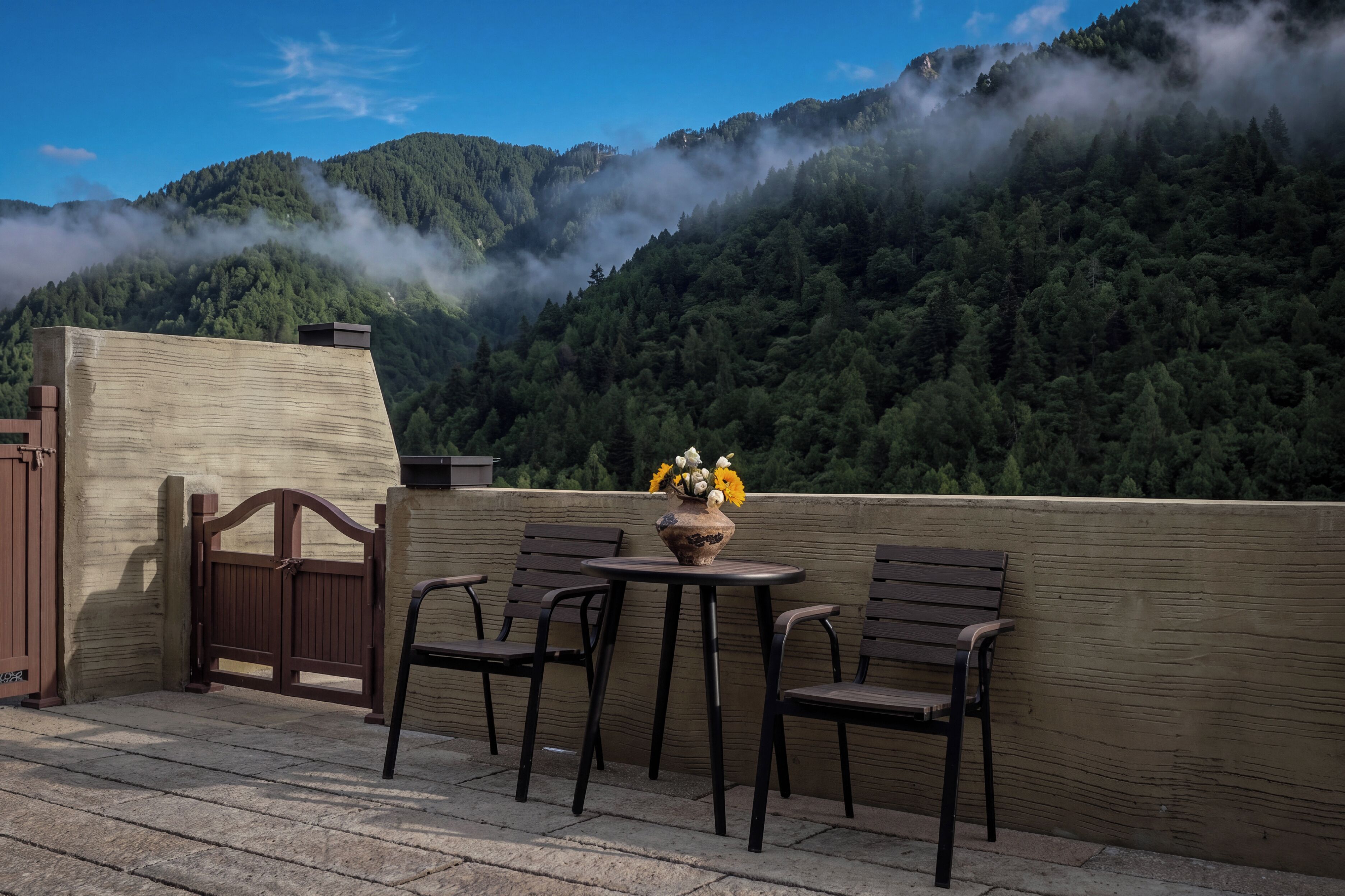 An outdoor seating area with a table and chairs, a vase with flowers, and a view of a forested mountain.