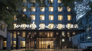 Entrada de hotel com letreiros iluminados, área de estar externa e um espelho d'água.