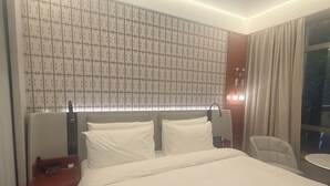 Quarto de hotel com uma cama grande, mesa de cabeceira com abajur, cadeira e janela com cortinas.