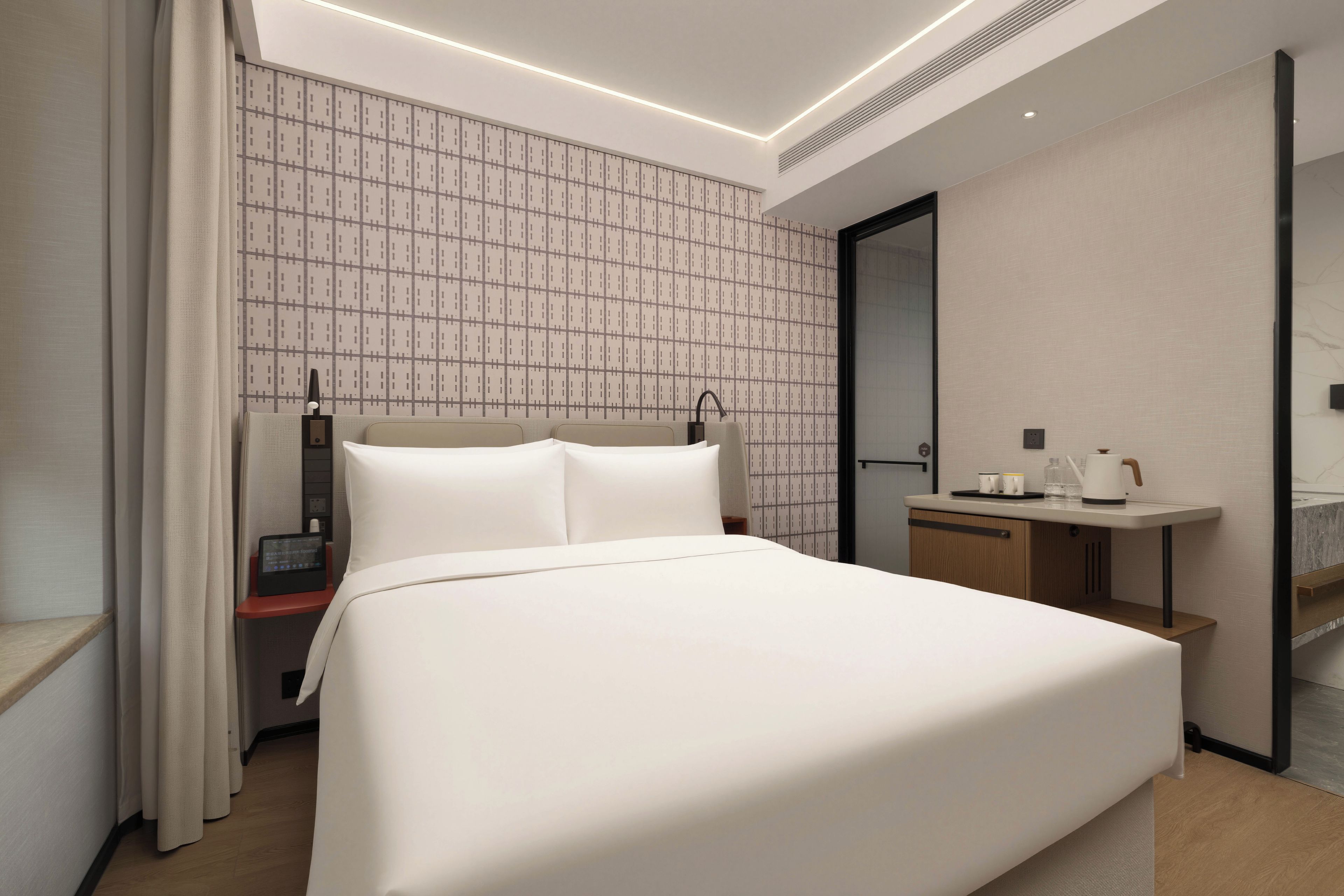 Ein modernes Hotelzimmer mit einem großen Bett, einem Nachttisch, einem integrierten Schreibtisch und einem Badezimmer mit Spiegel und Waschbecken.