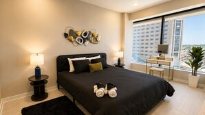 Een moderne slaapkamer met een groot bed, nachtlampen, een bureau en een uitzicht op een stadsgezicht door grote ramen.