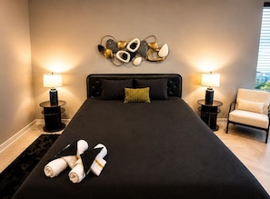 A hotel room with a large bed, two bedside lamps, a decorative wall piece, a chair, and a black rug. - Luxury 2BR | King Bed + Space to Gather (Birmingham)