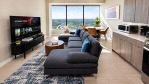 Uma sala de estar moderna com um sofá seccional, uma TV de tela plana e um espaço de refeições com vista para uma rodovia.
