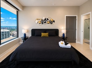 A modern bedroom with a large bed, a city view, a decorative wall piece, and a door leading to a shower. - Skyline Luxury | 2BR + King | Downtown BHM (Birmingham)