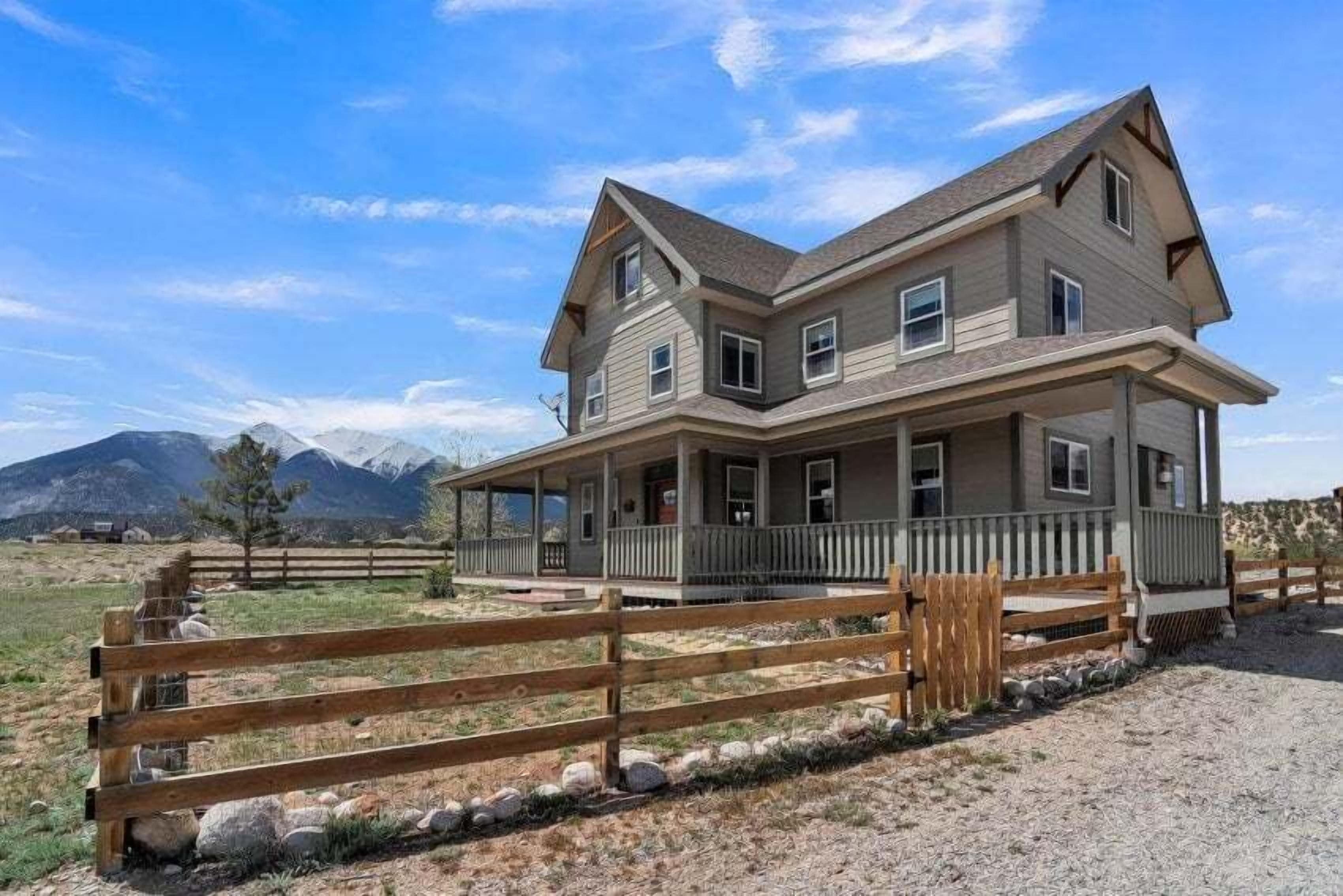 Une maison à deux étages avec un porche, une cour clôturée et des montagnes en arrière-plan.