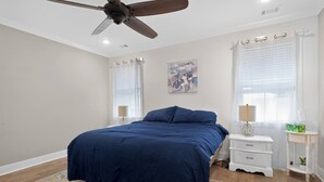 Ein Schlafzimmer mit einem Bett, einem Deckenventilator, einem Nachttisch, einer Lampe und einem Fenster mit Vorhängen.