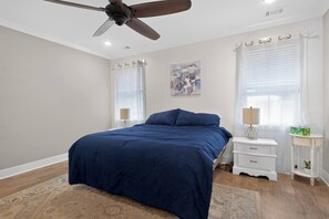 Ein Schlafzimmer mit einem Bett, einem Deckenventilator, einem Nachttisch, einer Lampe und einem Fenster mit Vorhängen.