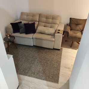 Uma sala de estar com um sofá bege, uma poltrona marrom escura e uma pequena mesa lateral com um vaso de flores.
