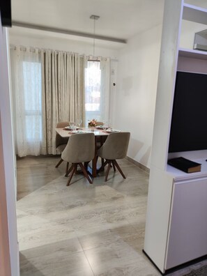Uma sala de jantar com mesa posta para quatro, cadeiras, um vaso com flores e uma televisão sobre um suporte.