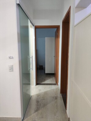 Corredor estreito com porta de vidro à esquerda, porta de madeira à direita e um banquinho ao fundo.